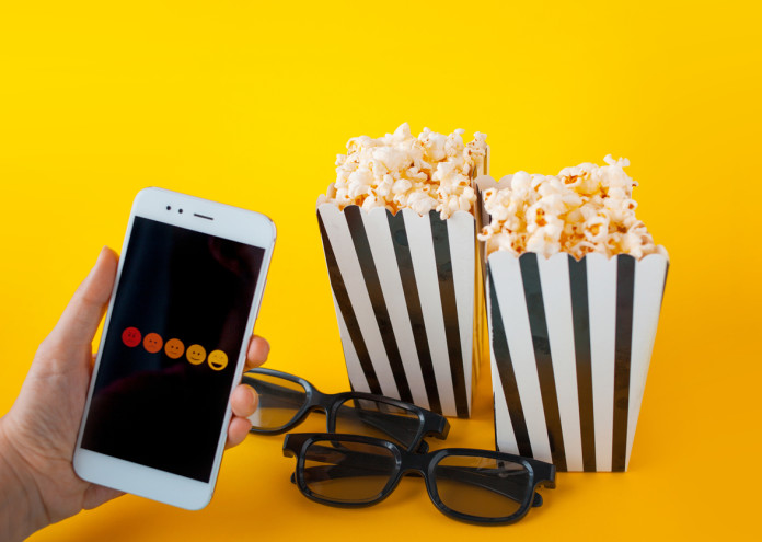  A hand is holding a smartphone in front of black-and-white paper boxes with popcorn and 3D glasses 